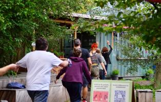 Eine Gruppe von fünf weißen jungen Leuten wurde von hinten fotografiert, wie sie in einer Reihe hintereinander her durch einen Garten laufen und Sportübungen mit ihren Armen machen. Links und rechts von ihnen sind Pflanzen und Bäume im Bild.